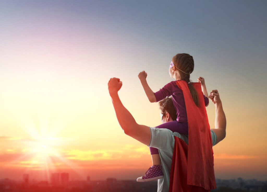 a father carrying his son on his shoulder wearing a mask and a red cape overlooking a sunset over city