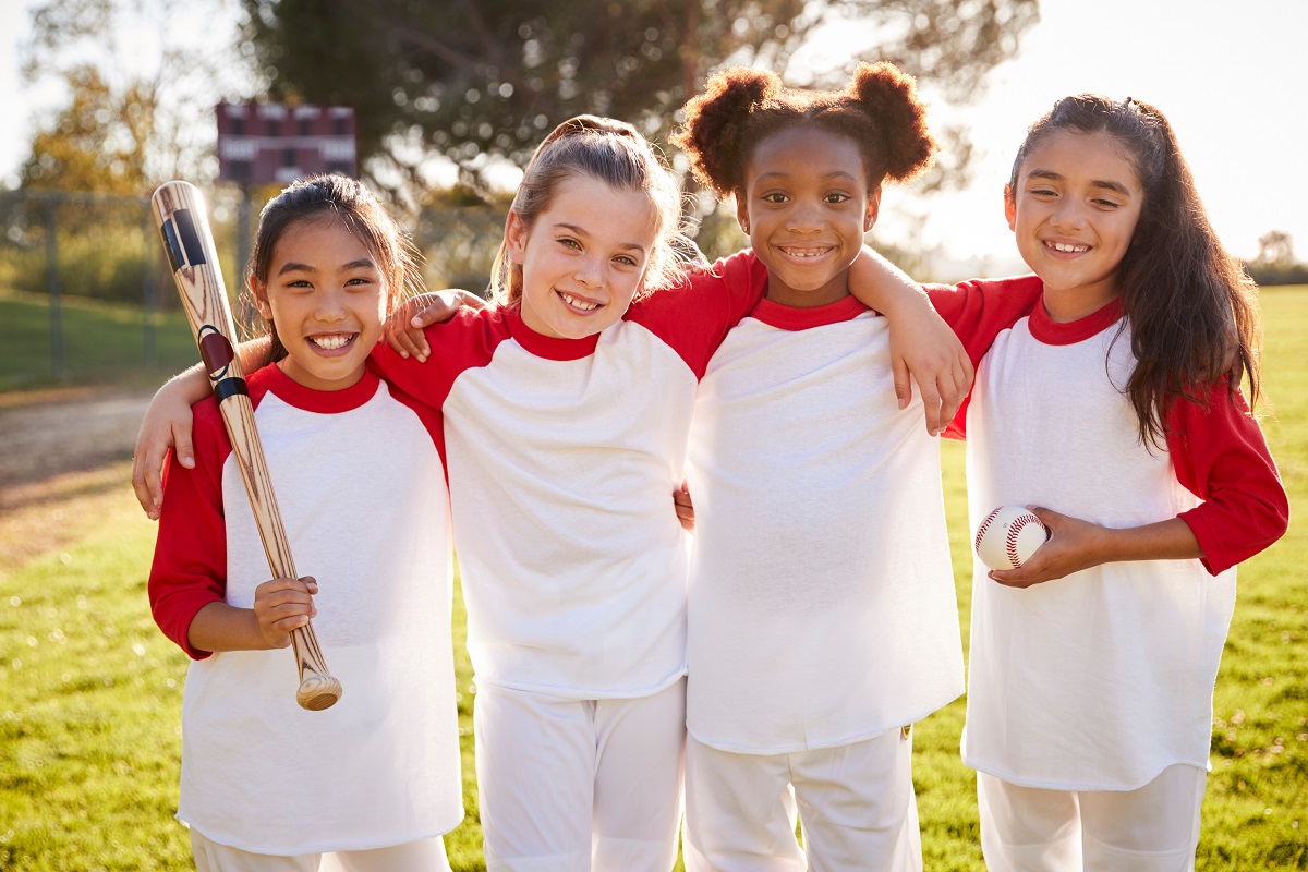 Young girls posing wearing baseball uniforms at a field.